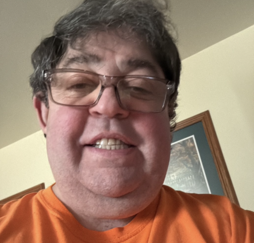 Portrait of James Allen Stanley, wearing a bright orange tee shirt and smiling