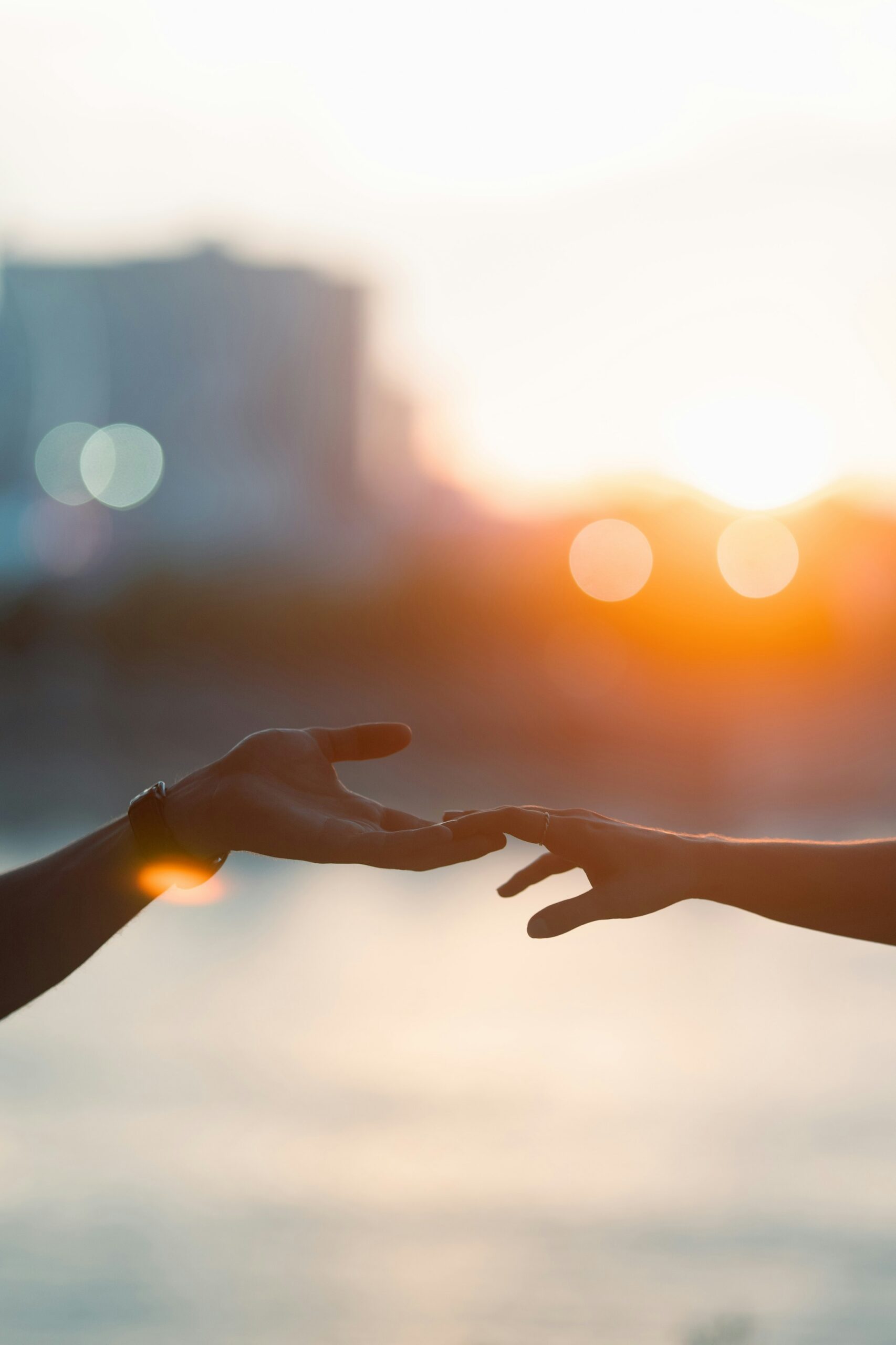 Two hands reaching toward each other at sunset over the water.