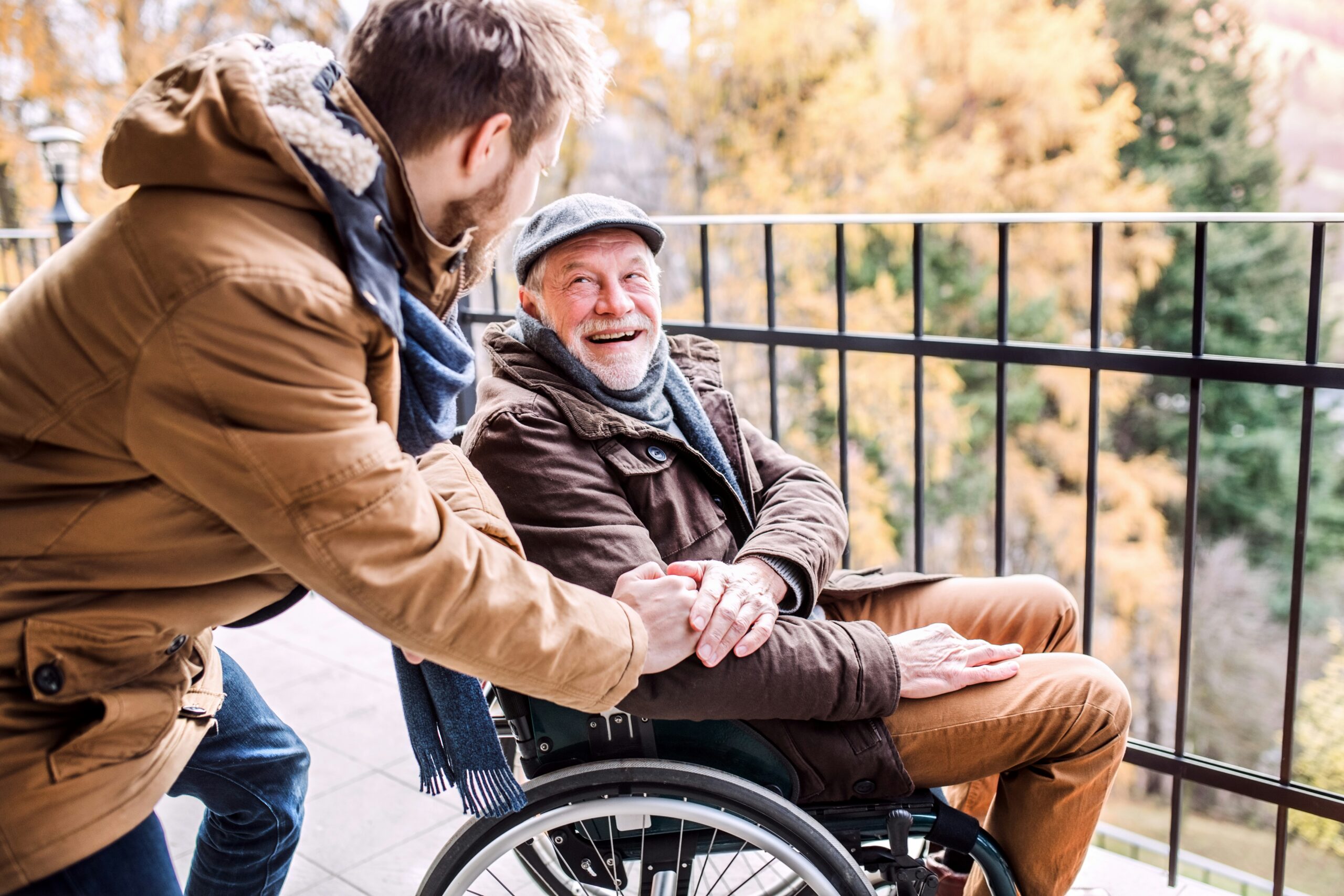 Older man with a white beard and wearing a cap sits in a wheelchair on a patio overlooking autumnal trees, smiling and holding the hand of a younger man with a beard.