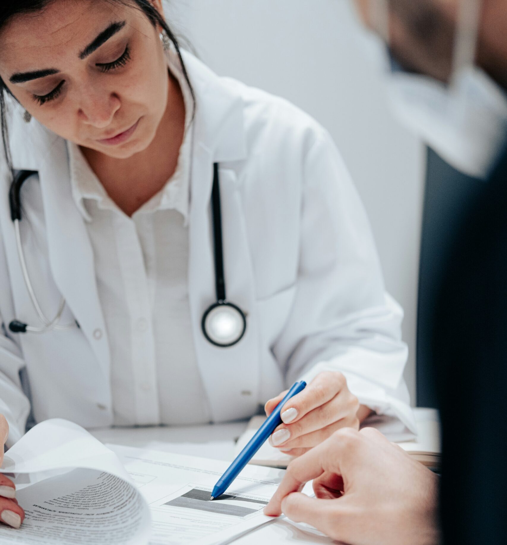 Female doctor with stethoscope around her neck going through paperwork on a clipboard with a male patient, who is blurred on the edge of the frame