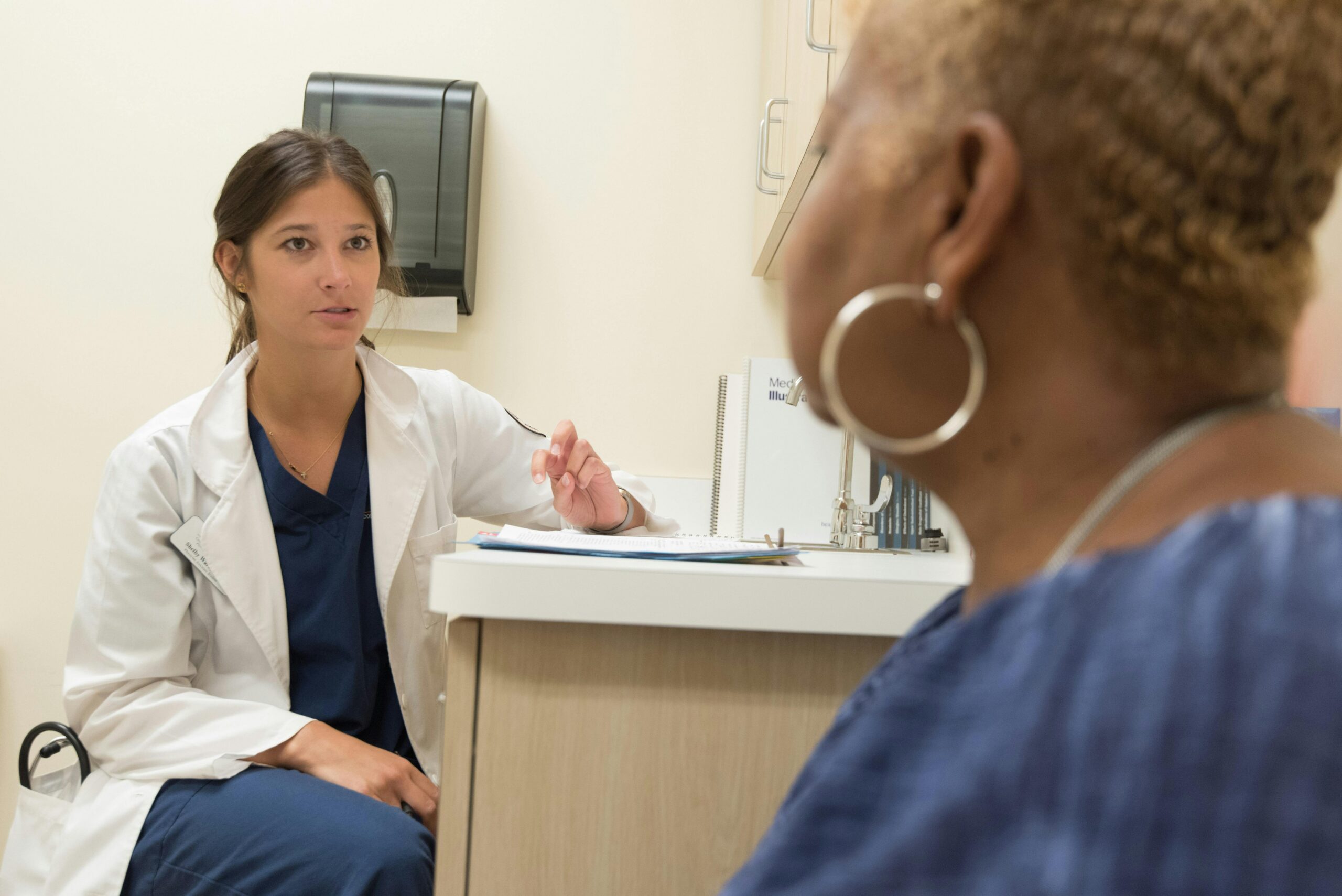 Female doctor wearing a physician coat sits down and has a conversation with a patient in doctor's office.