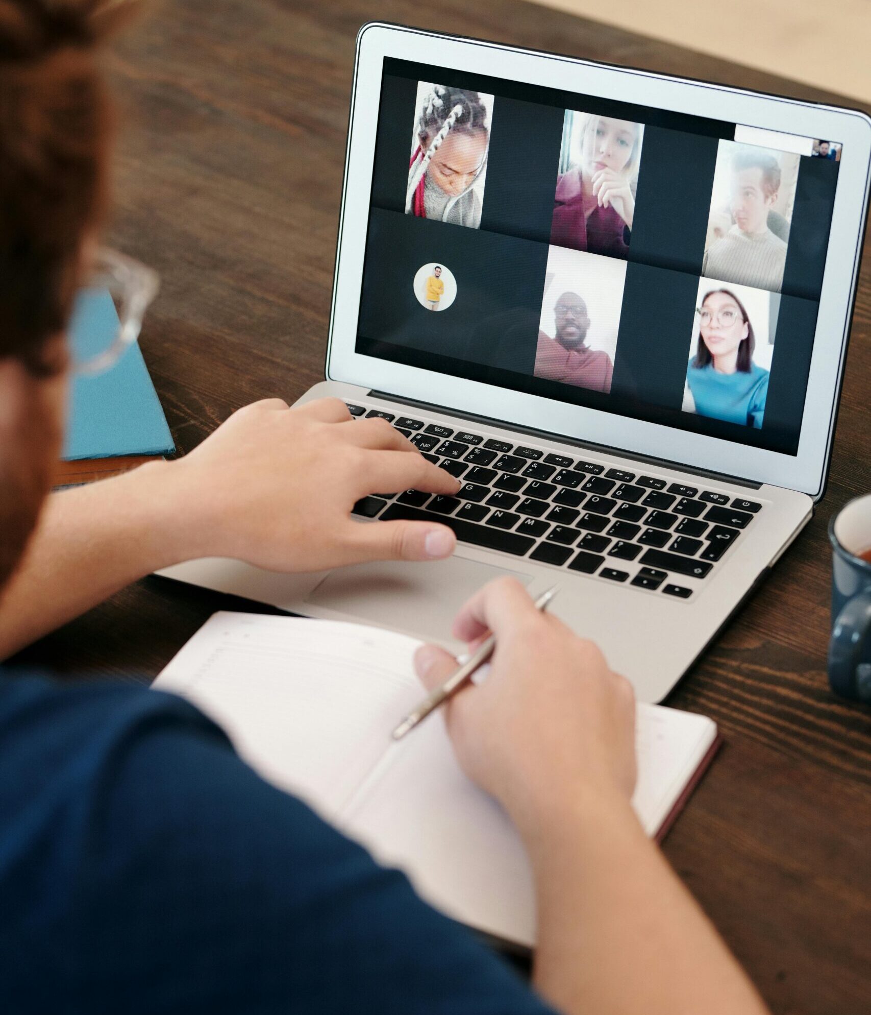 Person participating in a video call on a laptop while taking notes in a notebook at a desk.