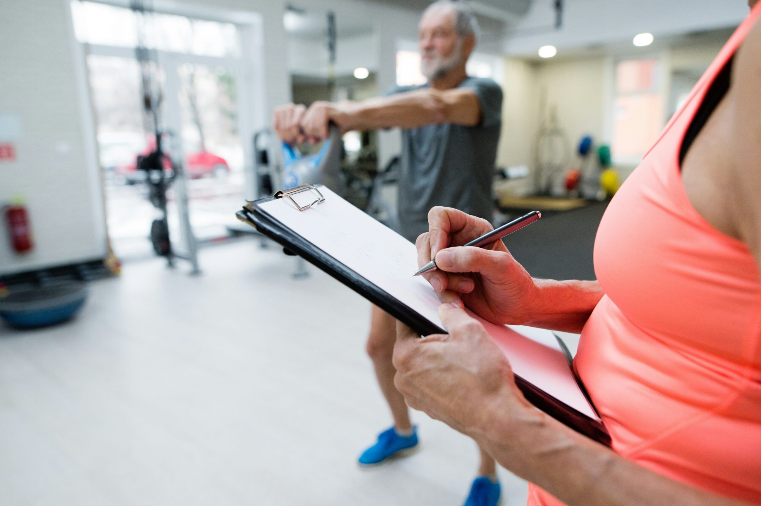 Older man exercising at a facility while a clinician takes notes on a clipboard in the foreground.