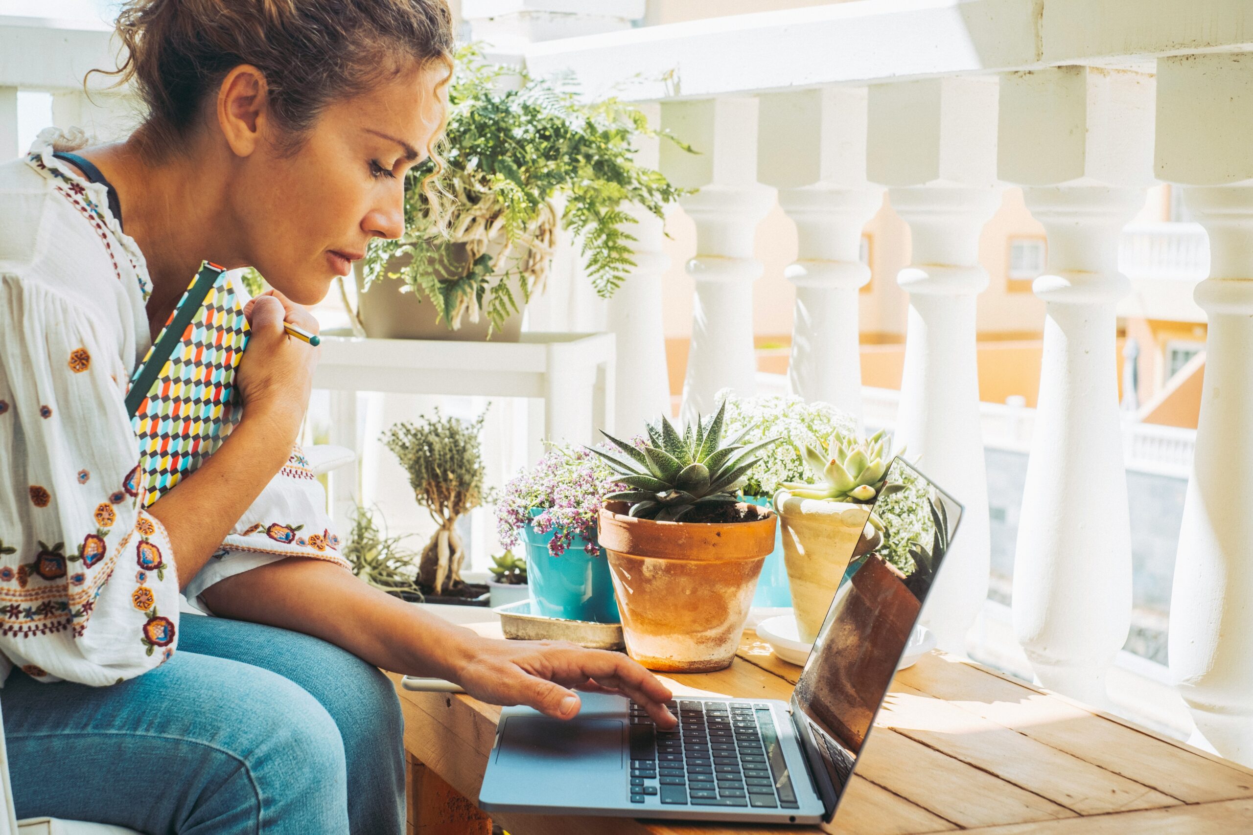Woman sitting on porch holding notebook while typing on a laptop