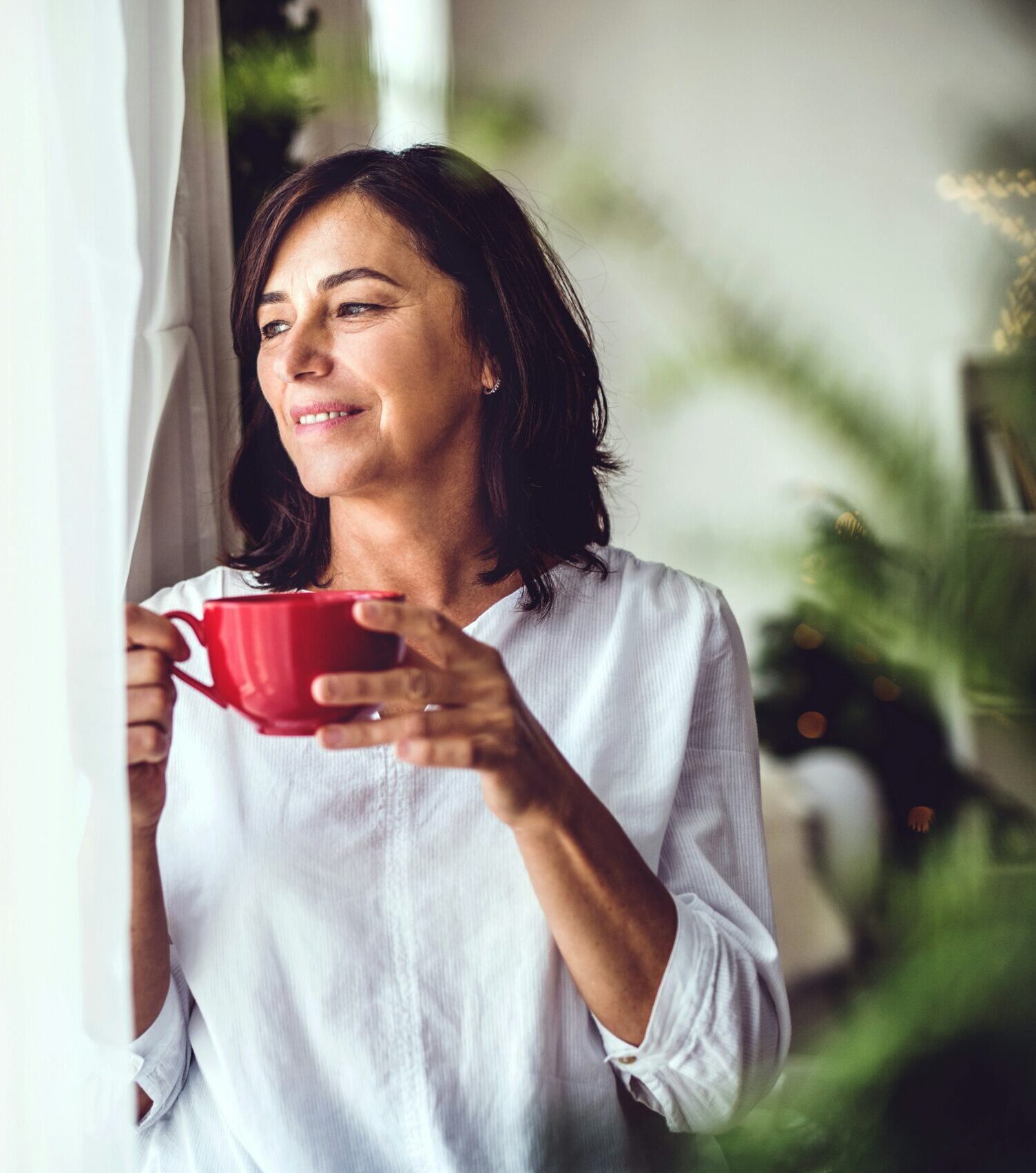 Woman looking out her window through sheer curtains holding red coffee mug.