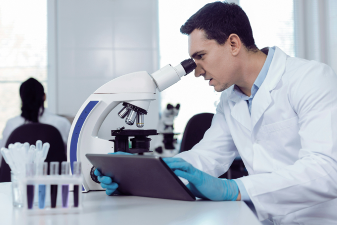 Researcher in a lab looking through microscope while holding a computer tablet