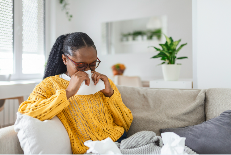 Woman sitting on couch blowing nose, with tissues scattered nearby